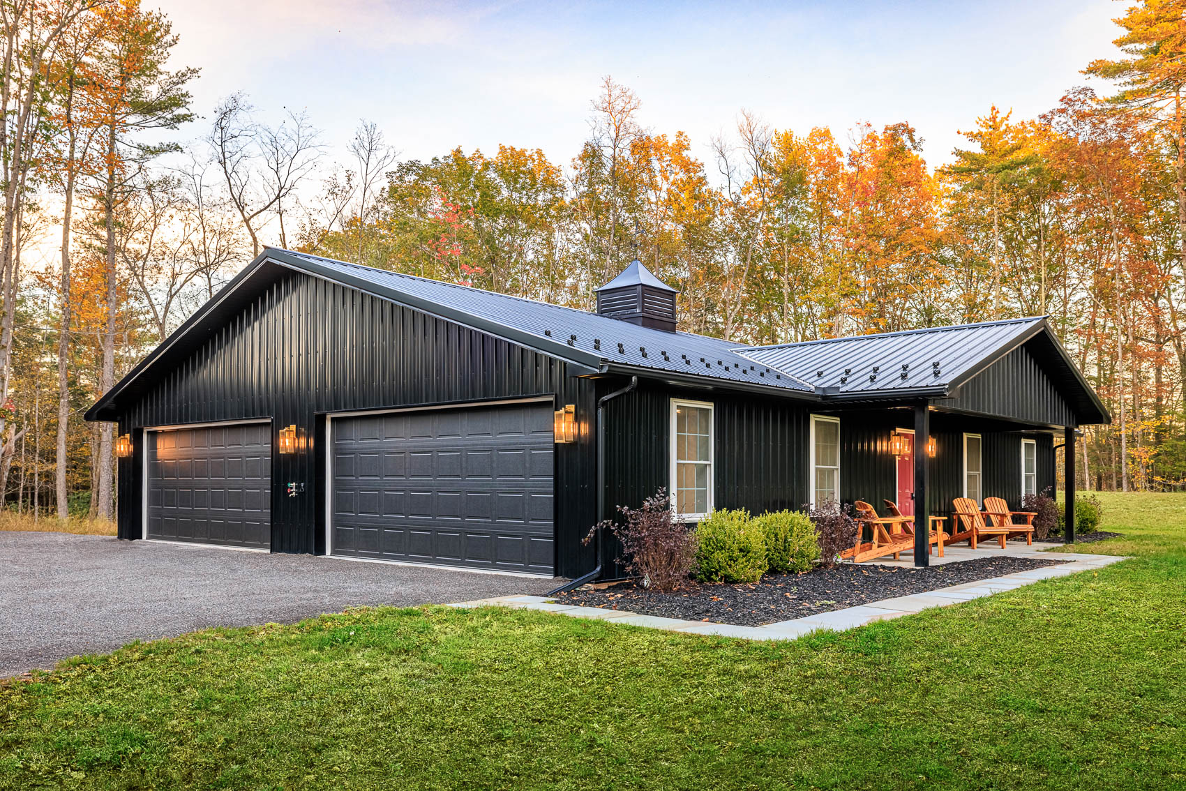 Black barndominium home with garage doors, porch seating, and autumn woods