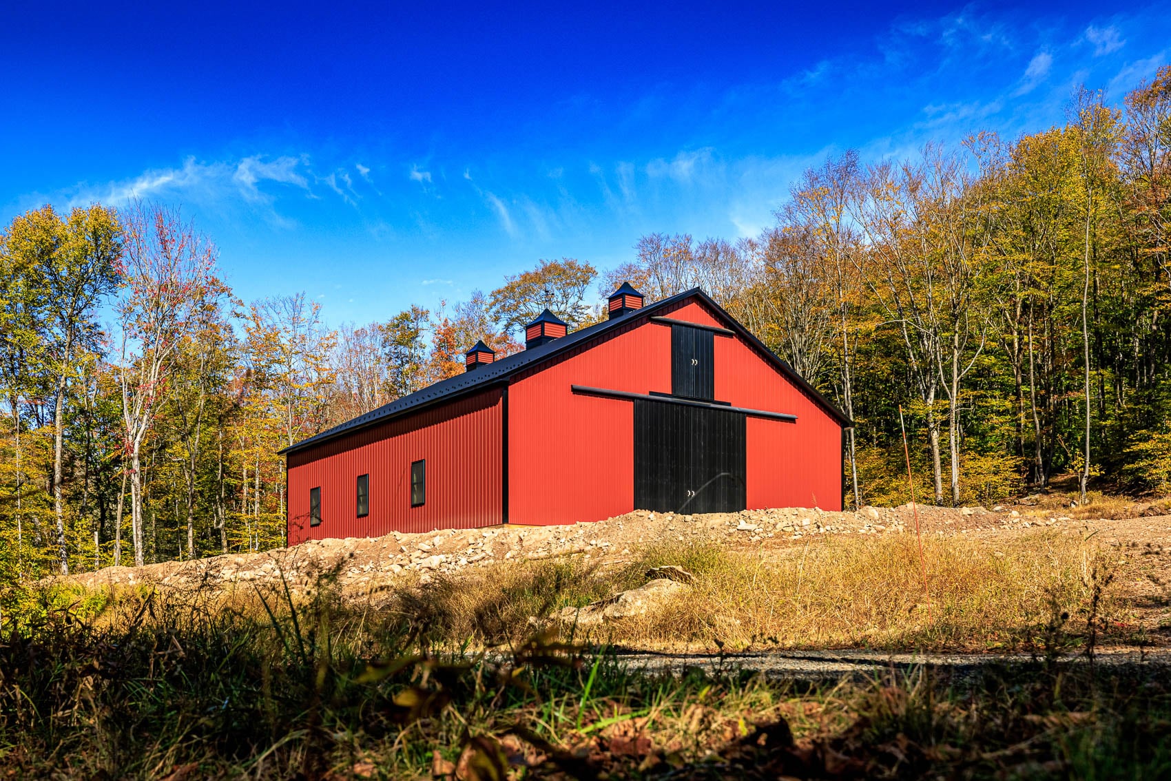 Low exterior view of red custom barn with black doors and autumn woods