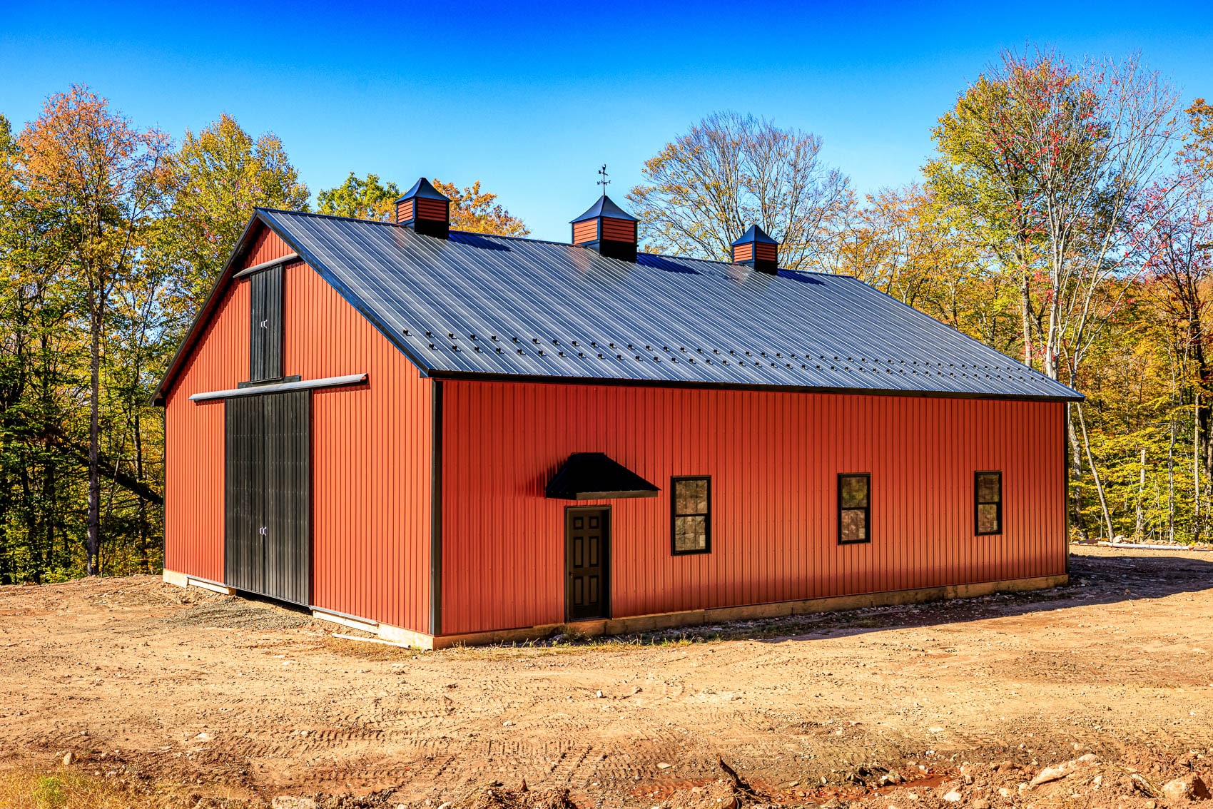 Red custom post-frame barn with black sliding doors, black roof, and cupolas